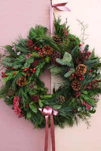 The Wild About Flowers Love Actually Winter Wreath, adorned with pinecones, red berries, green pine branches, and a pink ribbon bow, hangs beautifully on a light-colored door.