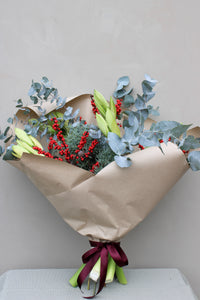The Mulled Wine Luxury Stems bouquet by Wild About Flowers is wrapped in brown paper with a maroon ribbon, featuring eucalyptus, light green lilies, red berries, and green foliage set against a plain light gray background.