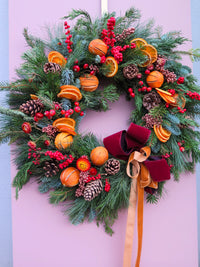 The Mulled Wine Wreath from Wild About Flowers features pinecones, dried orange slices, red berries, and a burgundy ribbon bow, beautifully set against a pinkish-purple background for a festive touch.