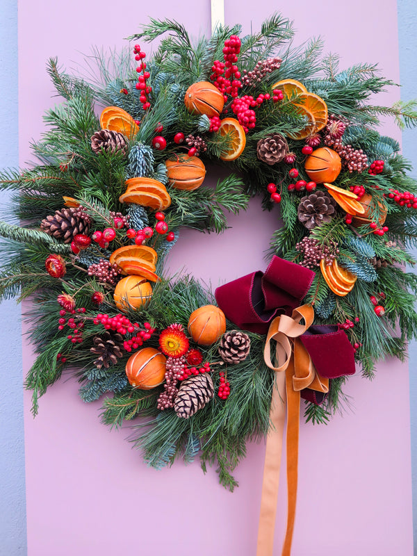 The Christmas Time Wreath from Wild About Flowers features pinecones, dried orange slices, red berries, and lush greenery. A velvet red bow completes the look as it elegantly decorates a pastel pink door, adding vibrant holiday décor perfect for embracing the festive tradition.