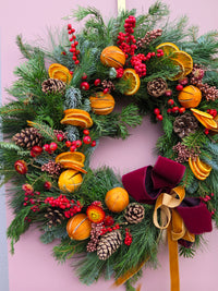 The Wild About Flowers Mulled Wine Wreath, with pine, pinecones, red berries, dried orange slices, and a red velvet bow with gold ribbon, makes a festive door decoration against a pale pink background.