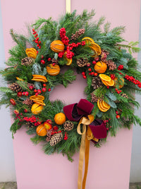 The Wild About Flowers Mulled Wine Wreath, featuring pine branches, cones, red berries, dried orange slices, orange ornaments, and a burgundy-gold ribbon, hangs as a festive door decoration on a pale pink background.