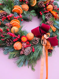 The Mulled Wine Wreath by Wild About Flowers features pinecones, dried orange slices, red berries, and a burgundy velvet ribbon with tan streamers—creating a festive touch for your door against a light pink background.