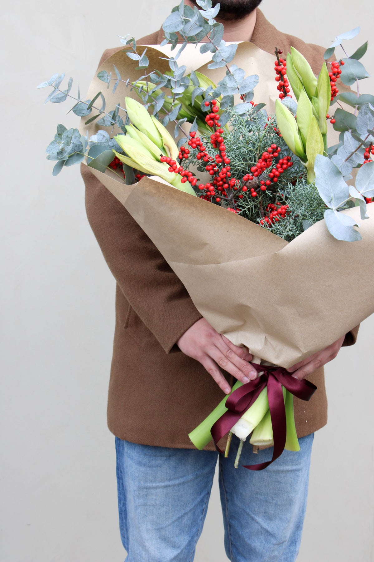 Wearing a brown coat and jeans, a person holds the "Driving Home for Christmas Stems" by Wild About Flowers, featuring lilies, red berries, eucalyptus, and greenery. The bouquet is wrapped in brown paper with a burgundy ribbon, beautifully contrasting the plain light-colored wall.