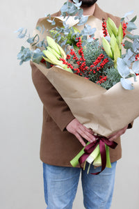 A person in a brown coat and blue jeans holds Wild About Flowers' Mulled Wine Luxury Stems—festive Christmas flowers with yellow lilies, red berries, eucalyptus, and greenery—wrapped in brown paper and tied with a dark ribbon.