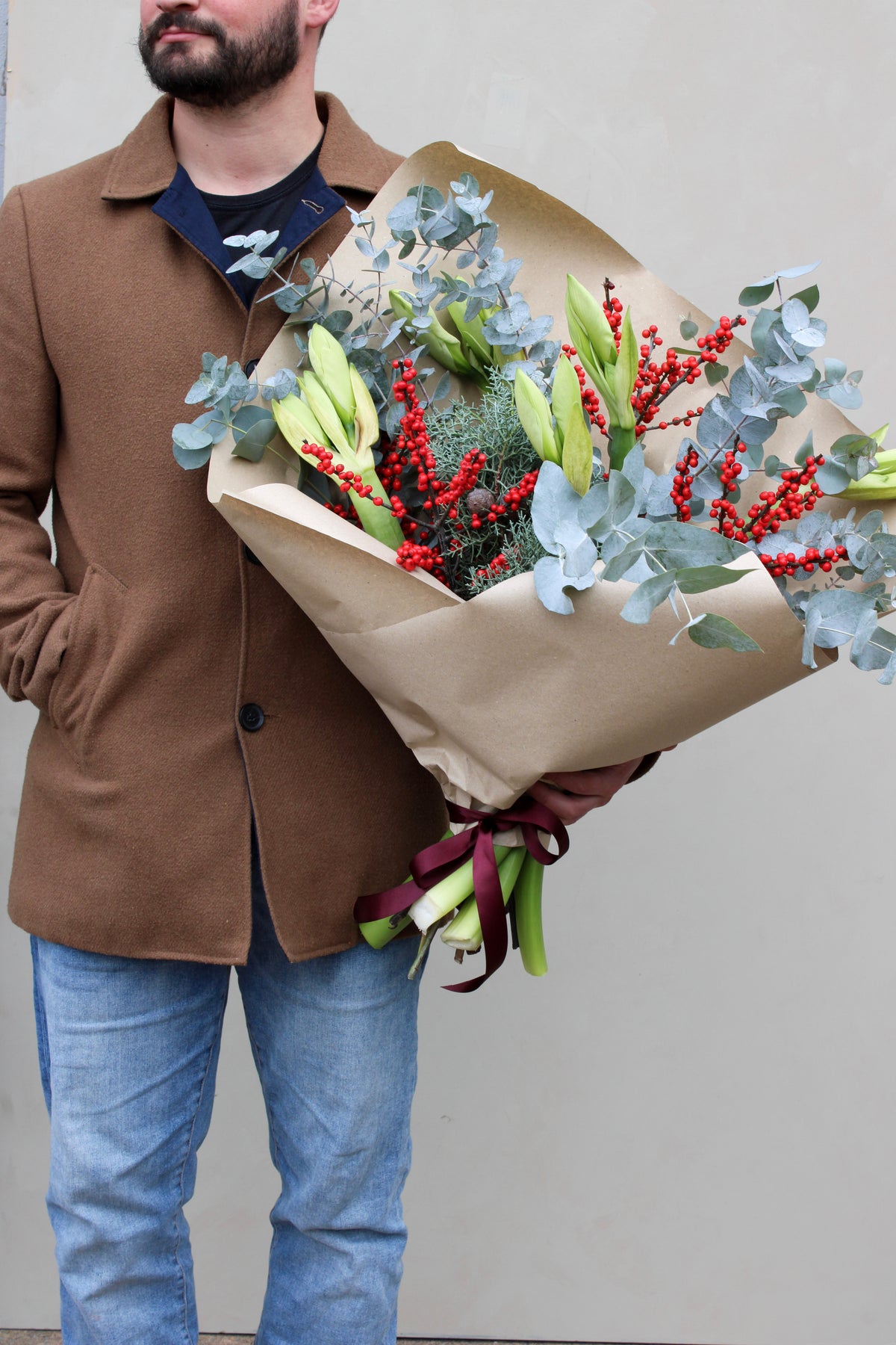 Against a plain light wall, a person in a brown coat and jeans holds Wild About Flowers' "Driving Home for Christmas Stems," a festive bouquet featuring white lilies, vibrant Amaryllis, red berries, and eucalyptus leaves, wrapped in brown paper with a burgundy ribbon.