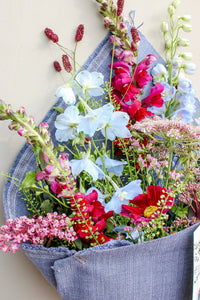 The Berry Cooler by Wild About Flowers features a colorful bouquet of wildflowers—light blue, red, pink, and white blooms with green stems—wrapped in upcycled denim for a charming and sustainable floral gift.