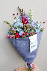 The Berry Cooler bouquet by Wild About Flowers features assorted wildflowers in pink, blue, and red, wrapped in upcycled denim with a pink ribbon and card—an eco-friendly gift set against a light-colored wall.