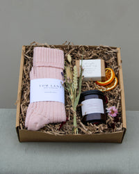 Introducing the Winter Warmers Gift Box by Wild About Flowers: a cozy set with pink knitted socks, a soothing candle, fragrant soap, and decorative dried flowers and orange slices, all beautifully arranged in shredded paper inside a brown cardboard box.
