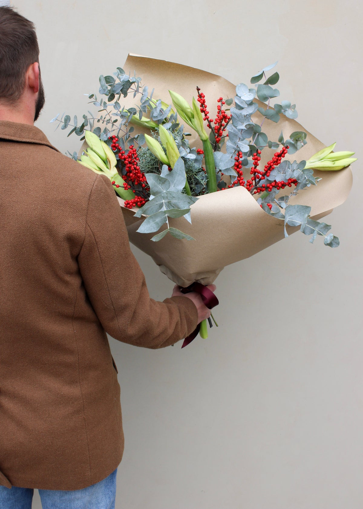 A person in a brown jacket holds the "Driving Home for Christmas Stems" bouquet by Wild About Flowers, beautifully wrapped in brown paper. This festive arrangement features green foliage, red berries, white lilies, and possibly elegant amaryllis blooms—ideal for the holiday season.