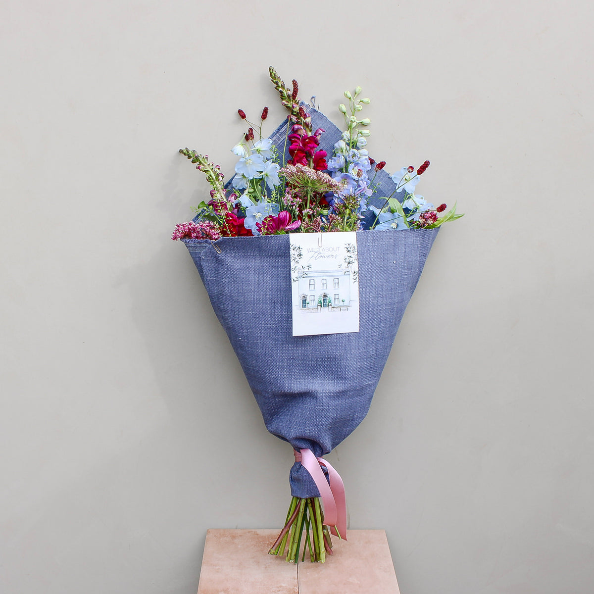 The Berry Cooler by Wild About Flowers features assorted flowers in an upcycled denim wrap, tied with a pink ribbon and a card, leaning upright against a light beige wall. A chic, sustainable floral gift.