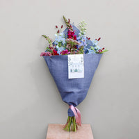 The Berry Cooler by Wild About Flowers features assorted flowers in an upcycled denim wrap, tied with a pink ribbon and a card, leaning upright against a light beige wall. A chic, sustainable floral gift.