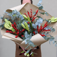 A person holds the Mulled Wine Luxury Stems bouquet by Wild About Flowers, wrapped in brown paper with green lily buds, eucalyptus, and red berries—a festive Christmas arrangement set against a plain background.