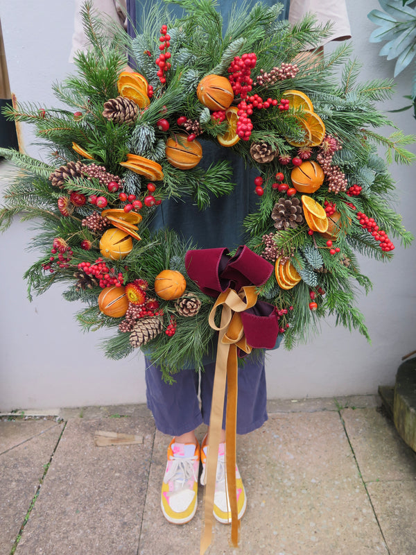 A person holding a Wild About Flowers' Christmas Time Wreath, crafted from green pine branches, pinecones, dried orange slices, red berries, and adorned with a maroon velvet bow featuring gold ribbons. Ideal for the festive season, the individual is wearing colorful sneakers and standing on a stone surface.