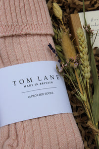 A close-up of pink alpaca bed socks with a "Tom Lane Made in Britain" label, nestled in a Winter Warmers Gift Box from Wild About Flowers. The scene includes dried flowers and wheat on crinkled brown paper, with a small envelope partially visible in the background.