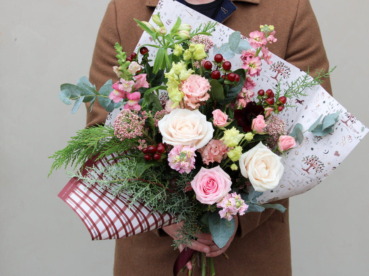 Someone wearing a brown coat is holding the Merry Little Christmas Bunch by Wild About Flowers, which showcases festive flowers such as pink and ivory roses, delicate pink blooms, red berries, and seasonal stems wrapped in decorative paper.