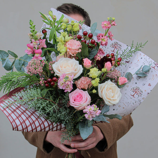 A person in a brown coat is holding the Merry Little Christmas Bunch by Wild About Flowers, showcasing festive pink, white, and green flowers. The bouquet is wrapped in decorative paper and features seasonal stems with roses, berries, and greenery that obscure the person's face.