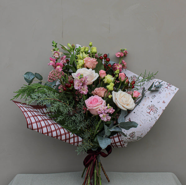 The Wild About Flowers' Merry Little Christmas Bunch displays seasonal stems of pink and white roses complemented by lush green foliage, all gracefully wrapped in plaid-patterned paper. This festive bouquet stands beautifully against a simple gray backdrop, capturing the essence of the holiday season.