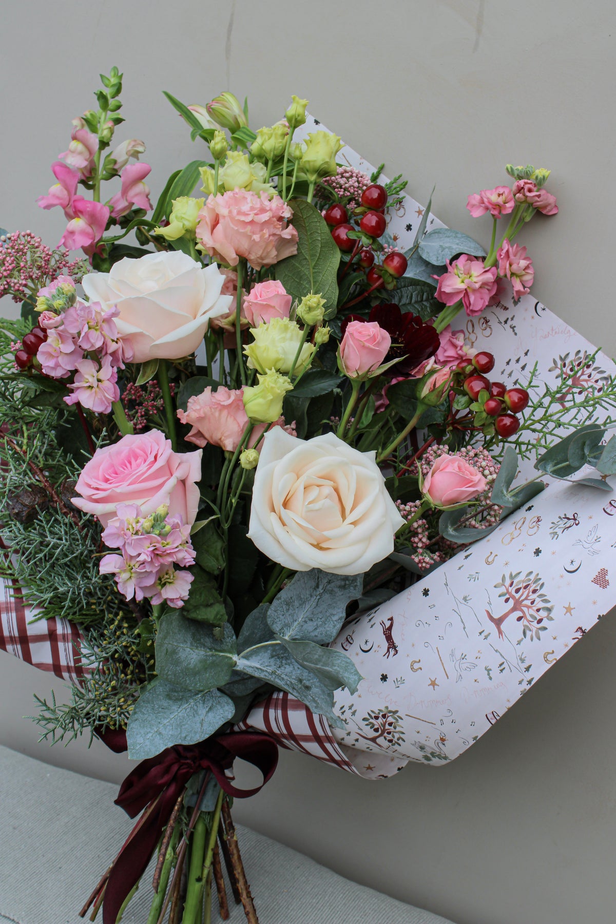 The Merry Little Christmas Bunch by Wild About Flowers features an exquisite combination of pink and white roses, carnations, and seasonal stems beautifully wrapped in decorative floral paper with a plaid ribbon. This arrangement includes lush green foliage and vibrant red berries set against a light gray background for a festive touch.