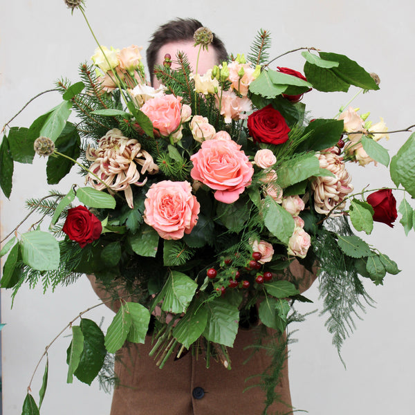 A person holding a large bouquet from Wild About Flowers, named Santa Baby Blooms, showcases pink and red roses along with assorted greenery and mixed flowers, partially obscuring their face against a neutral background.