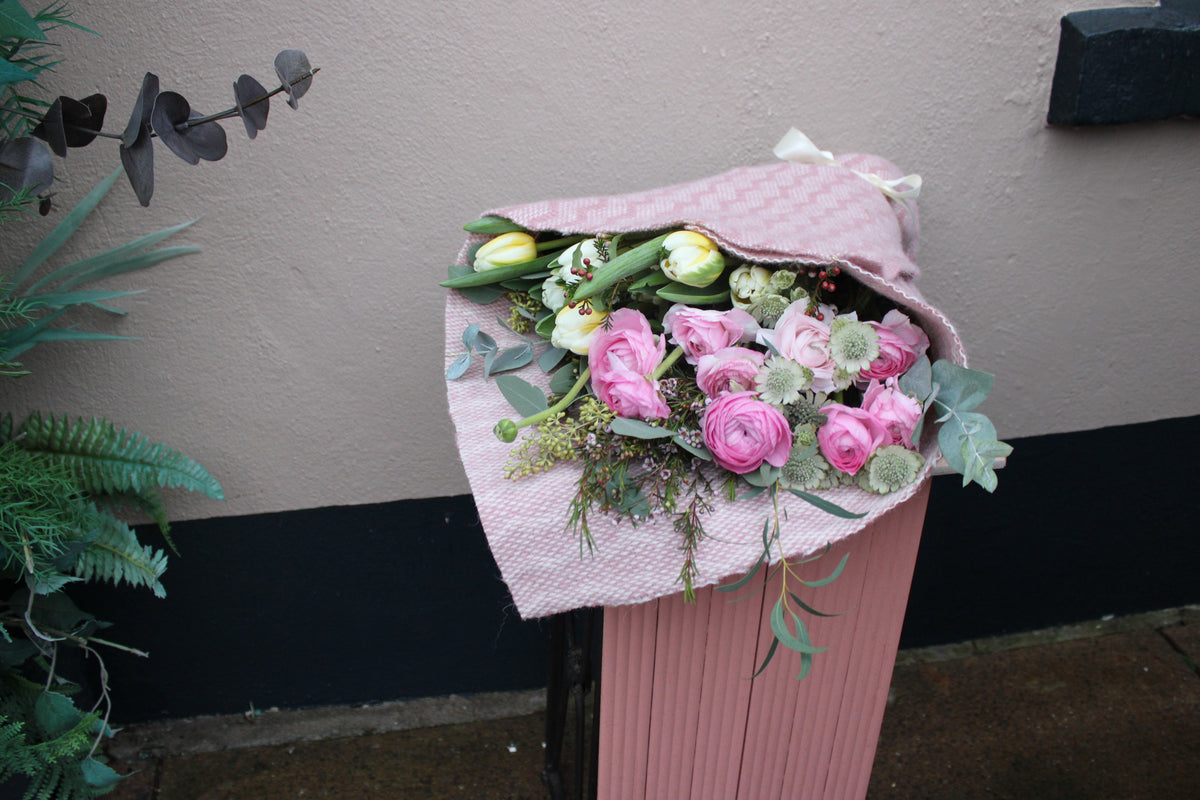 The 'Armful of Love' Valentines wrap by Wild About Flowers—featuring pink roses, white tulips, and greenery in textured paper—sits atop a pink trash bin against a beige-and-black wall.