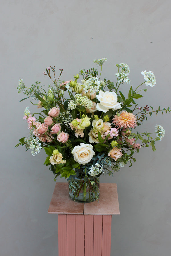 The "Beautiful Blooms" bouquet from Wild About Flowers features a stunning collection of assorted flowers, including elegant white and pink roses, arranged in a clear glass vase atop a pink pedestal against a plain gray background. The sophisticated florals, combined with greenery and small white blossoms, create a fresh and elegant display that exudes timeless design.