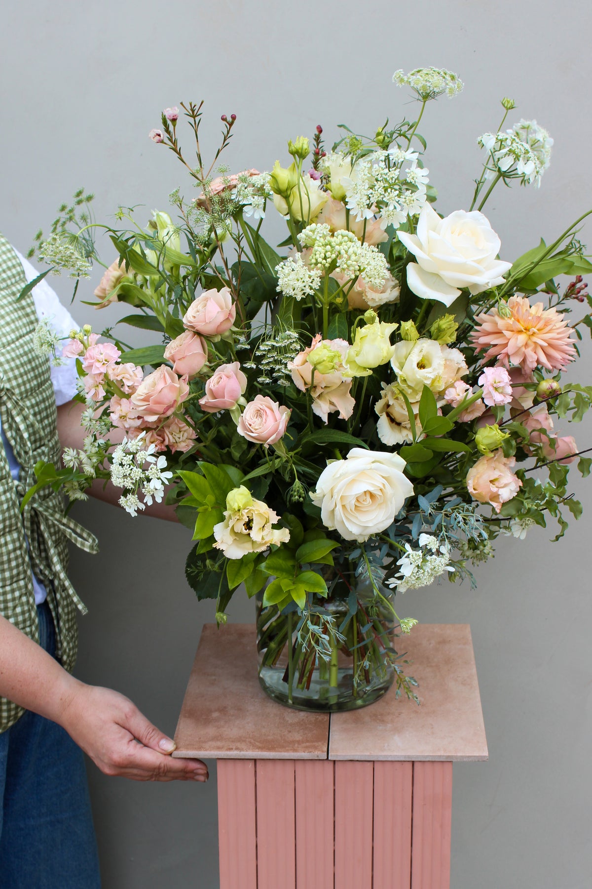 A person arranges a vase of Beautiful Blooms from Wild About Flowers on a pink pedestal. The bouquet features white, peach, and light pink roses interspersed with greenery and delicate white flowers. The person is wearing a green gingham vest and blue jeans. The background is plain gray, highlighting the timeless design of the arrangement.