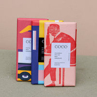 Three vibrant artisan chocolate bars from "Wild About Flowers" stand upright, featuring abstract wrappers: a red one with a person illustration, a blue and black one, and a pink, yellow, and blue one with geometric designs. This sustainable trio reads "Trio of Chocolate.