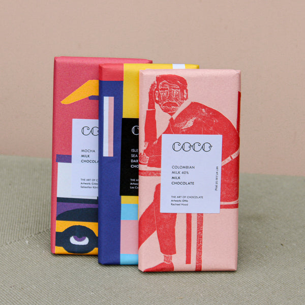 Three vibrant artisan chocolate bars from "Wild About Flowers" stand upright, featuring abstract wrappers: a red one with a person illustration, a blue and black one, and a pink, yellow, and blue one with geometric designs. This sustainable trio reads "Trio of Chocolate.