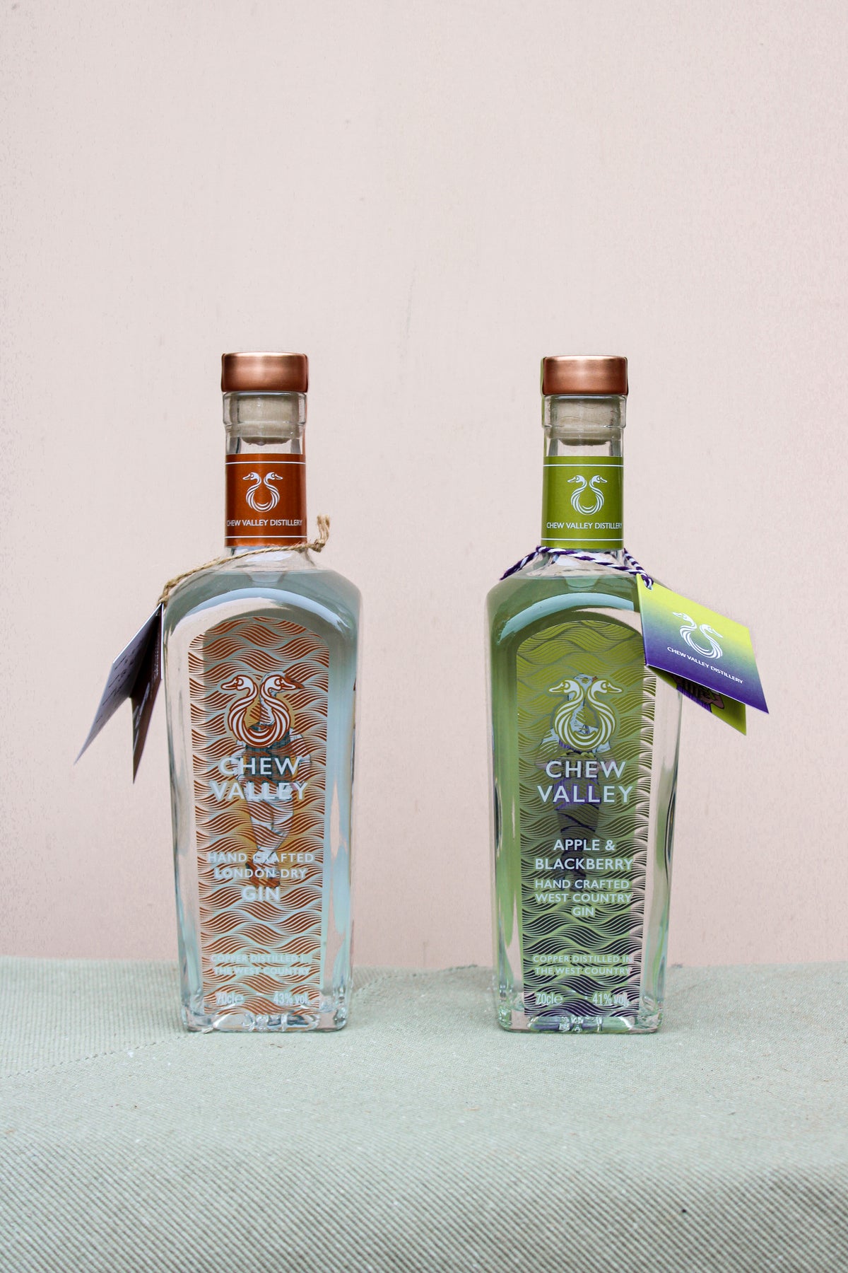 Two bottles of Chew Valley Gin by Wild About Flowers are on display: London Dry Gin with a copper cap and Apple & Blackberry Gin with a green cap. Crafted from locally sourced ingredients, they are elegantly set against a light background on a textured surface.