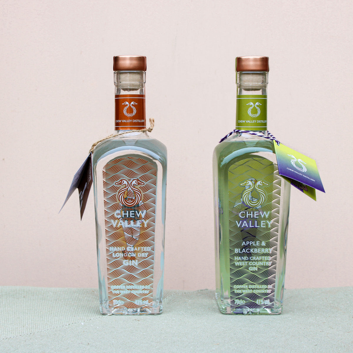 Two bottles of Wild About Flowers' award-winning Chew Valley Gin sit on a light green surface against a beige backdrop. The left bottle has a copper cap, the right features an apple and blackberry label with a green cap. Both handcrafted in Somerset, they have tags on their necks.