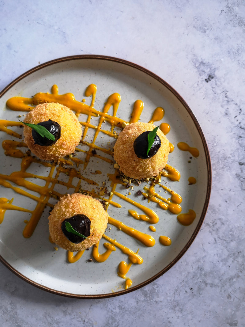 A marble plate features three crumb-coated appetizers from Supper Club with The Cooking Flea, topped with black garnishes and small green leaves, drizzled with a yellow sauce made from seasonal produce.