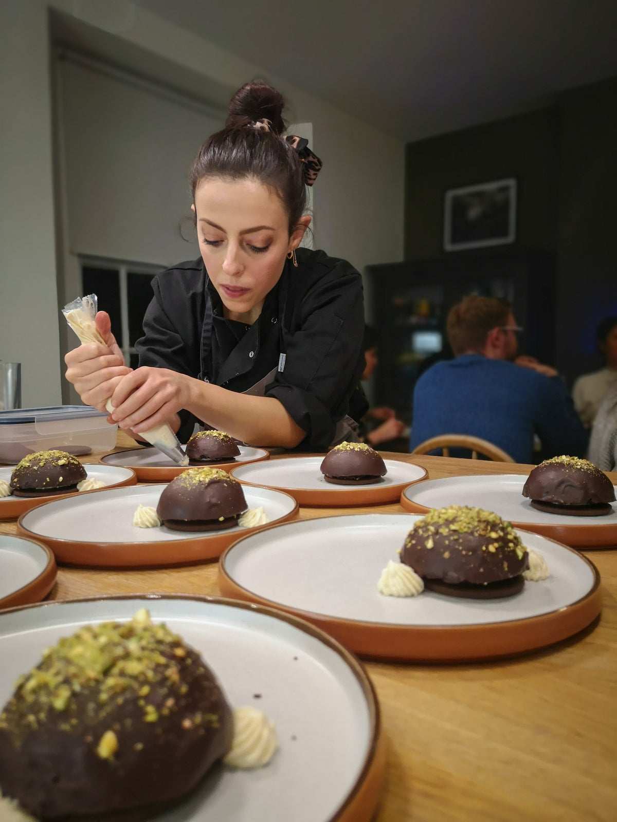 Amidst the warmly lit room, a woman in a black chef's coat prepares chocolate domes garnished with crushed pistachios on white plates during the exclusive "Supper Club with The Cooking Flea" event, crafting an engaging culinary experience as others enjoy the scene.