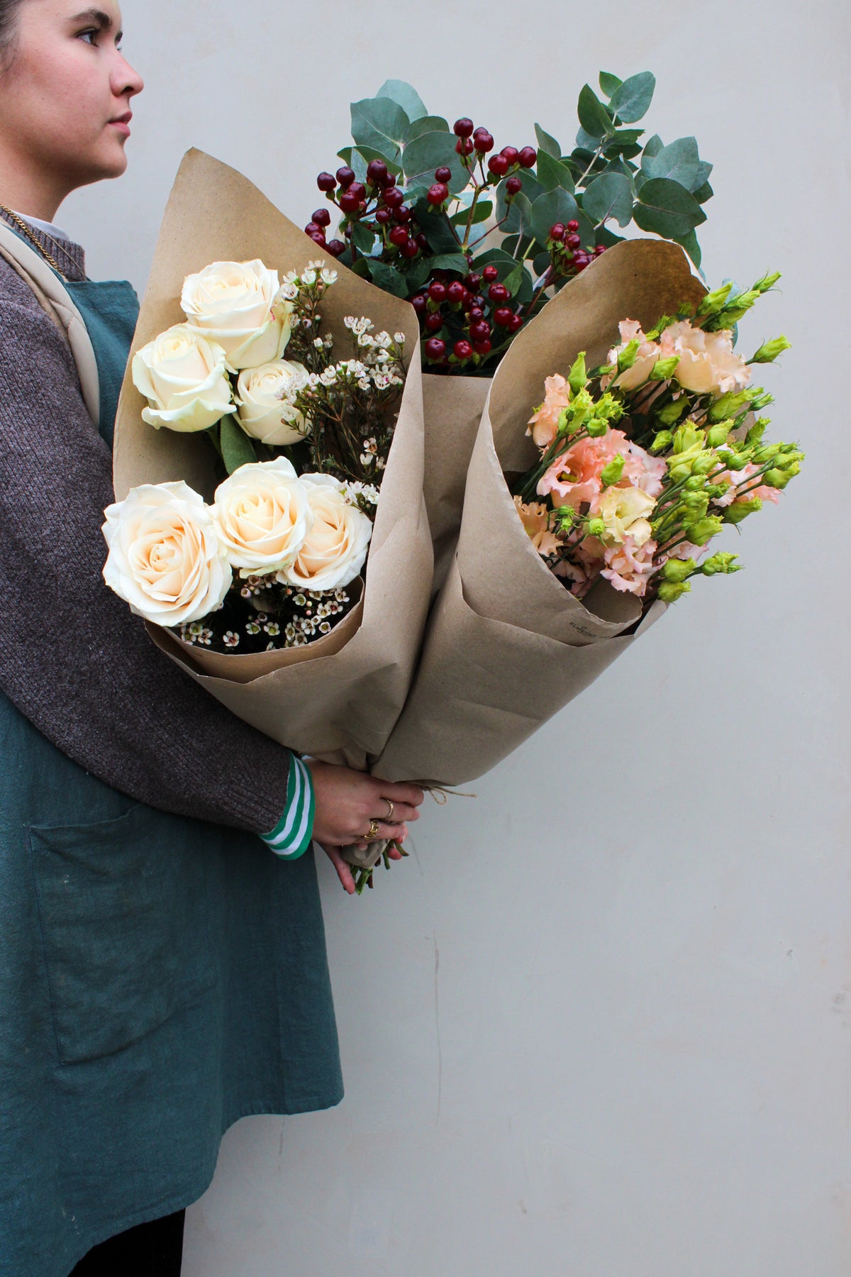 A person in a teal apron displays three "House Full of Flowers" bouquets by Wild About Flowers, wrapped in brown paper. The arrangements feature seasonal blooms like white roses, greenery, red berries, and light peach flowers against a plain background.