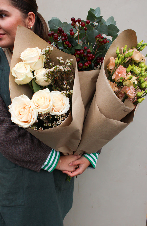 A person wearing a green apron over a striped shirt holds three "House Full of Flowers" bouquets by Wild About Flowers, showcasing exquisite artistry with white roses, small pink flowers, red berries, and lush green foliage wrapped in brown paper.