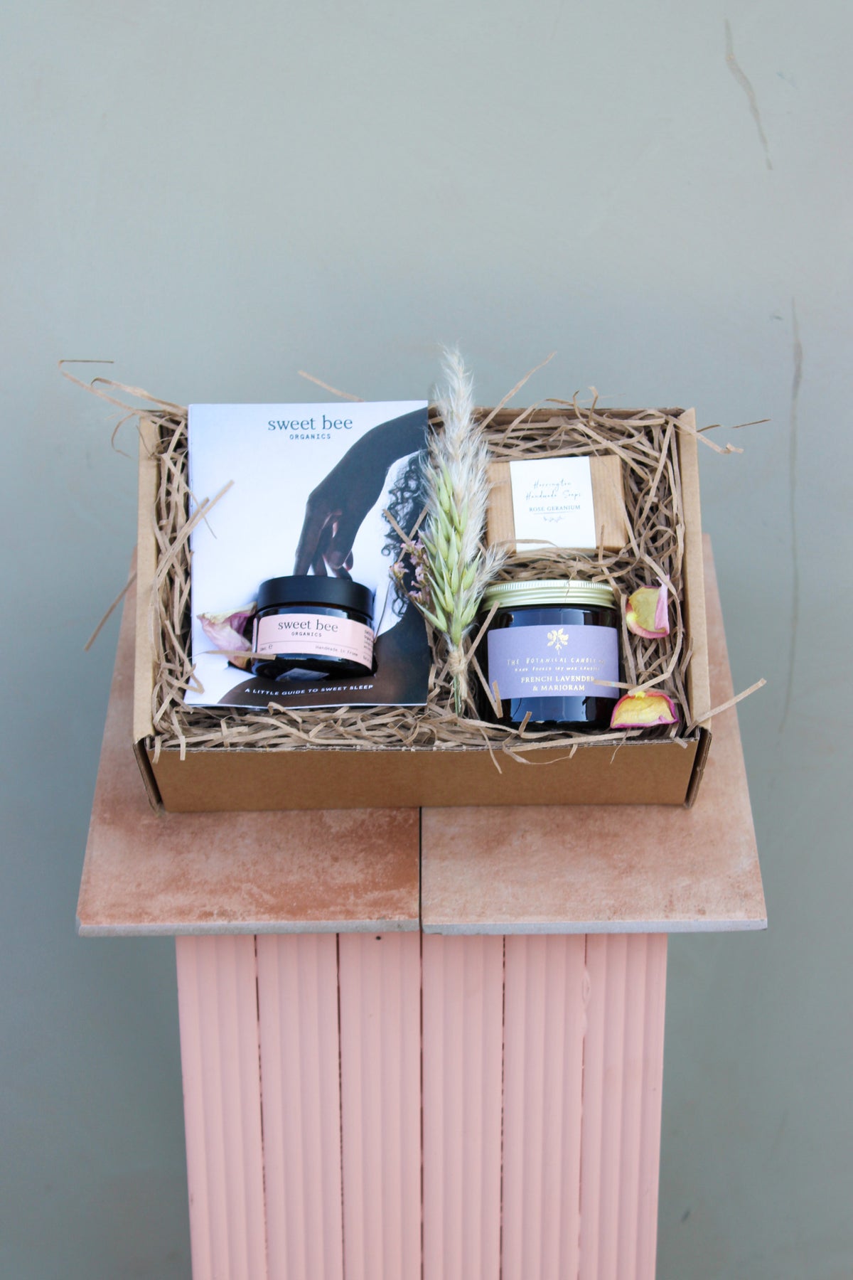 The Wild About Flowers Mother's Day Gift Box features all-natural beauty items, including a candle, Sweet Bee Magnesium Butter, a cork-lidded jar, booklet, and dried flowers—all elegantly arranged in a charming brown box on a pink pedestal with straw lining.