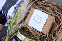 The Mother's Day Gift Box by Wild About Flowers includes a "Rose Geranium" bar of Harrington Handmade Soaps in crinkled paper, dried wheat stalks, a jar lid with a gold pattern, and Sweet Bee Magnesium Butter for soothing delight.