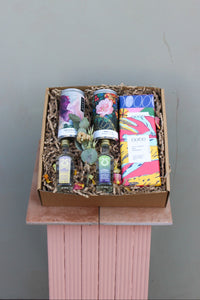 The Mother's Day Hamper by Wild About Flowers, displayed on a pink pedestal, includes two cans, two small bottles, decorative plants, and colorful chocolate bars. The artistic packaging and shredded paper filler create an elegant and delightful presentation.