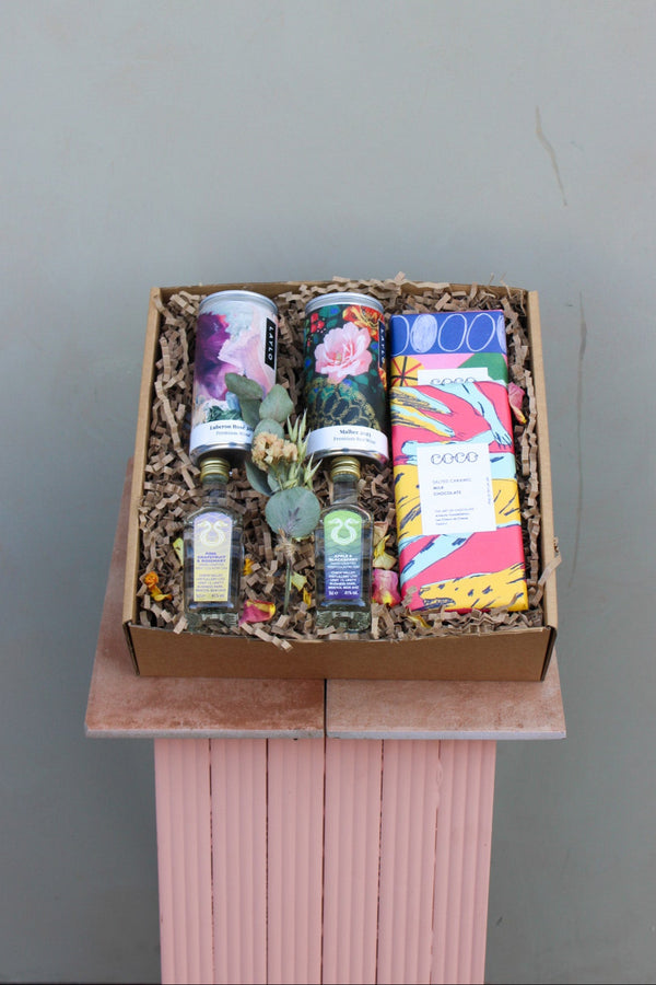 The Mother's Day Hamper by Wild About Flowers, displayed on a pink pedestal, includes two cans, two small bottles, decorative plants, and colorful chocolate bars. The artistic packaging and shredded paper filler create an elegant and delightful presentation.