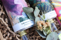 Close-up of wine bottles in crinkled packaging. One, labeled "Luberon Rosé 2021 Premium Wine," adorned with abstract art, embodies exquisite wines. Dried eucalyptus leaves and small flowers enhance the charm, ideal for Wild About Flowers' Mother's Day Hamper.