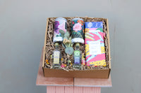 The Mother's Day Hamper by Wild About Flowers includes two botanical gins, a floral wrapped item, and three patterned packages in a decorative paper box with an eucalyptus leaf. It's elegantly presented on a pink pedestal against a plain backdrop.