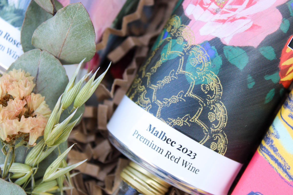 A close-up of a Mother's Day Hamper by Wild About Flowers features a premium Malbec 2023 red wine bottle with a colorful, floral label. Dried flowers and greenery are artfully arranged beside the bottle on a bed of crinkled paper shavings.