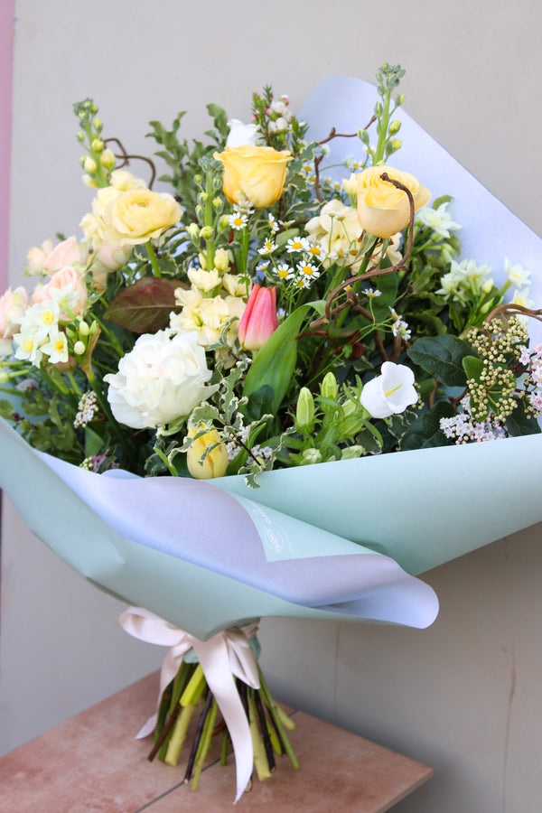 The Wild Thing bouquet by Wild About Flowers, wrapped in light blue paper with yellow roses, white flowers, daisies, a pink tulip, green leaves, and stems, elegantly graces a wooden surface against a neutral background—ideal for Mother's Day gifting.