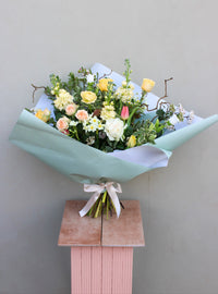 The Wild Thing bouquet by Wild About Flowers, featuring yellow, peach, and white roses with greenery and delicate blooms, is wrapped in light blue paper with a white ribbon. It elegantly sits on a pink pedestal against a light gray background—perfect for Mother's Day with local Keynsham delivery available.