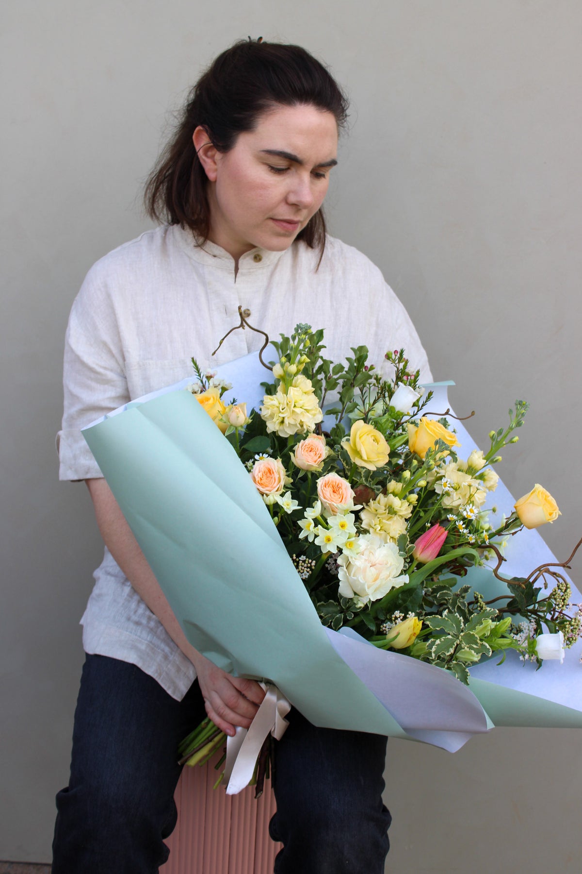 Cradling a stunning "Wild Thing" by Wild About Flowers, the person dons a light shirt and dark pants. This Hand-Tied Design bouquet in light blue paper showcases yellow/light pink roses, white flowers, lush greenery; perfect as a Mother's Day gift from your Keynsham florist against a neutral backdrop.