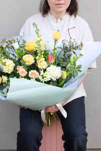 A person in a white shirt and dark pants holds the Wild Thing bouquet by Wild About Flowers, featuring mixed blooms with yellow roses, pink tulips, and white blossoms in light blue wrapping. Ideal for Mother's Day from your local Keynsham florist. A plain light wall is in the background.