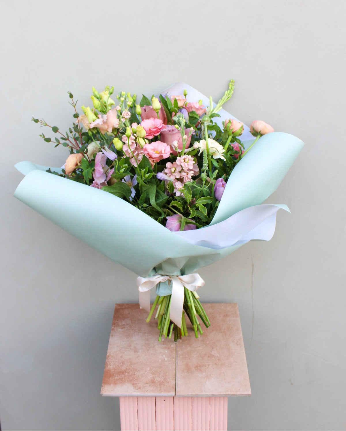 The "Secret Garden" by Wild About Flowers is a luxury bouquet with pink, peach, and green tones. Elegantly wrapped in light blue paper and tied with a white ribbon, it is artistically displayed on a light pink pedestal against a gray background.