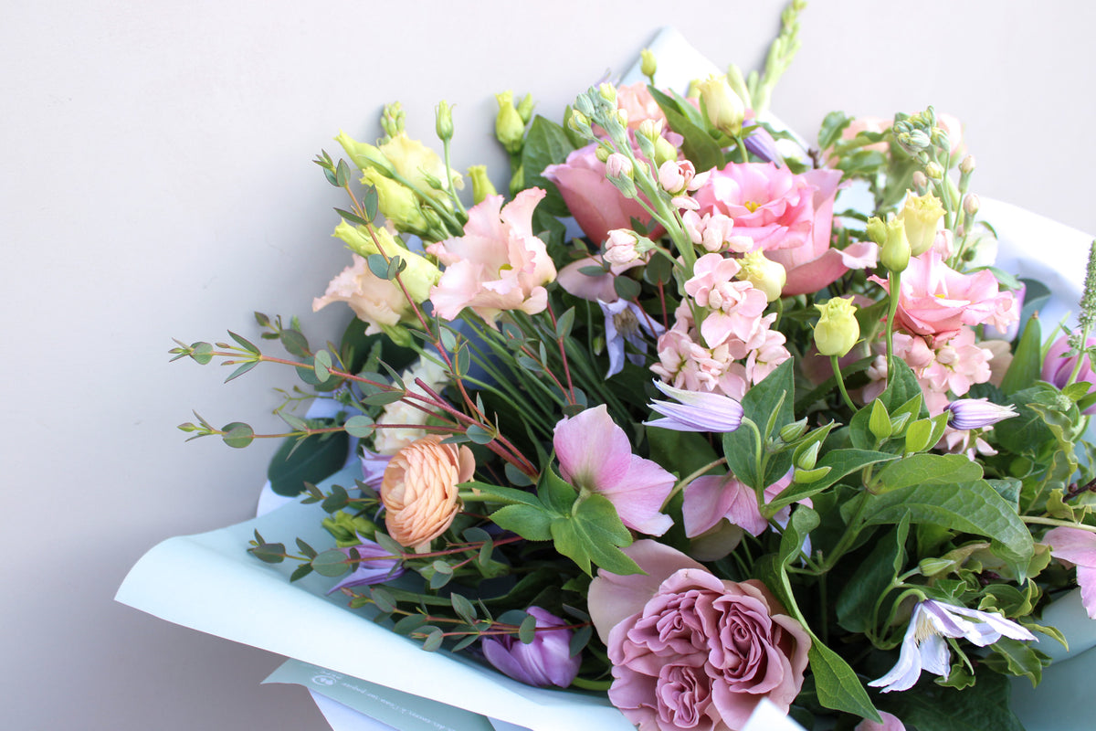 The "Secret Garden" bouquet by Wild About Flowers features a luxurious mix of pink roses, peach ranunculus, and greenery elegantly wrapped in light blue paper. This hand-tied arrangement showcases delicate blooms artfully displayed on a plain white background.