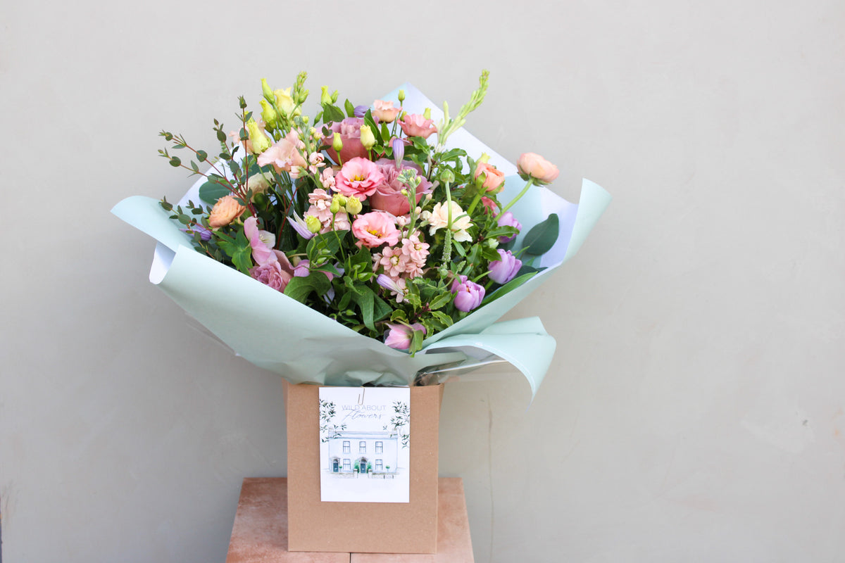 Wild About Flowers' Secret Garden features a luxurious bouquet of pink and purple flowers with greenery, beautifully wrapped in light blue and set in a brown box. A small card with a house drawing completes the arrangement, all set against a plain off-white background.