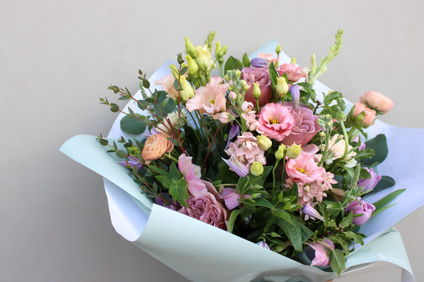 The "Secret Garden" by Wild About Flowers is a luxurious spring bouquet, showcasing pink and peach roses, lisianthus, and lush greenery in a hand-tied arrangement. It's elegantly wrapped in pale blue paper against a light gray backdrop.